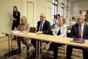 Ludzie siedzą w ławkach. Ich twarze skierowane są na wprost.