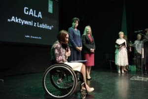 Na scenie są kobiety. Kobieta na wózku inwalidzkim ma na kolanach teczkę a w rękach mikrofon. Na prezentacji za nimi jest napis: “ Gala. Aktywni z Lublina”.