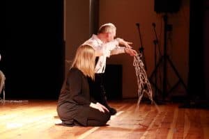 Na środku scenie klęczą kobieta i mężczyzna. Mężczyzna trzyma w ręku sieć a drugą wygina. Kobieta obok niego ma głowę skierowaną ku niemu. Są oni bokiem do obiektywu.