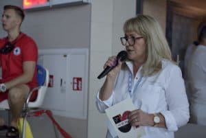 Kobieta przemawia do mikrofonu. W ręce trzyma broszurę.