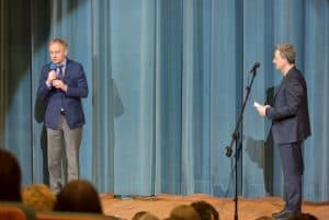 Mężczyźni stoją na scenie. Jeden trzyma w dłoni mikrofon. Drugi stoi przy mikrofonie stojącym i ma twarz zwróconą w strone drugiego meżczyzny.