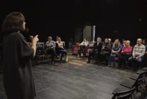 Kobieta stojąc na środku wskazuje palcem na publiczność. Widzowie siedzą naprzeciwko i po jej lewej stronie. Po prawej stronie rząd krzeseł jest pusty.