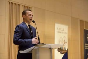 Mężczyzna w garniturze stoi na mównicy i przemawia do mikrofonu.