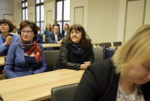 ludzie siedzą przy stołach. Ich twarze skierowane są na wprost.