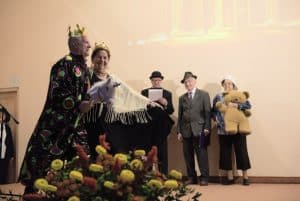 Na scenie stoi grupa starszych osób. Po lewej stronie mężczyzna jest przebrany za króla, ma koronę i głowę konia na kiju. Obok jest kobieta z korona na głowie. Z tyłu stoją dwaj mężczyźni i kobieta z dużym misiem.