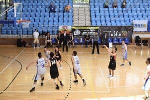 Mężczyźni grają na boisku. Po prawej stronie stoi mężczyzna z wyciągniętymi rękami do góry. W powietrzu leci piłka.