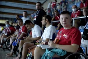 Mężczyźni na wózkach stoją w jednej linii. Chłopak najbliżej obiektywu patrzy na niego.