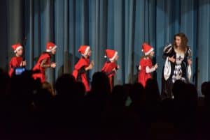 Kobieta w sukni jest na scenie. Po jej lewej stronie rzędę idą dzieci przebrane za mikołaje.