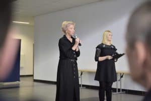 Kobieta stoi na środku pokoju i trzyma mikrofon w dłoni. Obok niej stoi kobieta, która trzyma teczkę w dłoni.