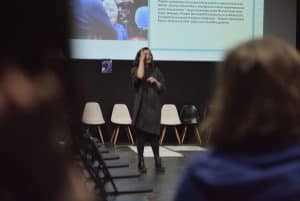 Kobieta stoi na środku zgromadzenia. Prawą dłoń ma skierowaną ku górze. Za nią wyświetlana jest prezentacja.