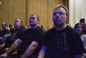 Osoby siedzą na widowni w rzędach. Jeden z mężczyzn patrzy wprost w obiektyw.