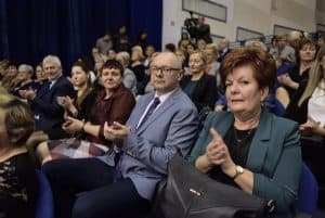 Osoby na widowni siedzą w rzędach blisko siebie. Wykonują oni gest klaskania. Dwie mkobety i mężczyzna patrzą w stronę obiektywu.