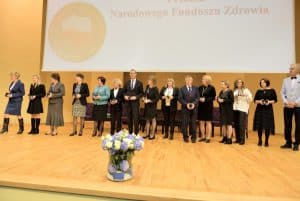 Grupa osób stoi na scenie. Wszyscy trzymają statuetki w rękach. Na nimi napis “ Narodowego Funduszu Zdrowia”.