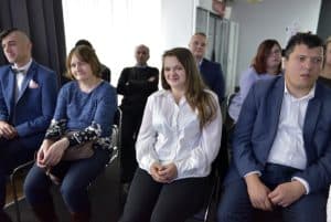 Ludzie siedzą na krzesłach. Niektórzy się uśmiechają. Twarze są zwrócone przed siebie.