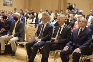 Grupa osób na widowni siedzi na krzesłach w rzędach. Wszyscy patrzą w jednym kierunku.