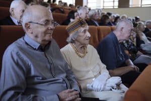 Duża grupa osób siedzi na widowni. Są to głównie osoby starsze. Wszyscy patrzą przed siebie.