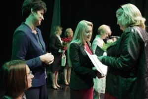 Na scenie jest grupa osób. Kobieta trzyma otwartą teczkę w kierunku stojącej przed nią kobiety.