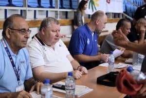 Czwórka mężczyzn w średnim wieku siedzi przy stole. Przed nimi dokumenty i woda.