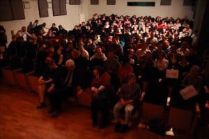 Na widowni siedzi duża grupa osób. Niektórzy siedzą w półcieniu.