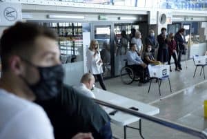 Kobieta przemawia do mikrofonu. Obok niej stoją ludzie lub siedzą na wózkach inwalidzkich. Po lewej stronie jest mężczyzna w maseczce który ma głowę odwróconą tak, że widać jego profil w obiektywie.