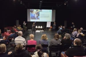 Jedna z pań stoi na środku sali, druga- przy tablicy. Kobieta z publiczności, trzyma mikrofon i stoi przy swoim krześle. W tle wyświetlana jest prezentacja.