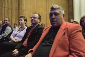 Osoby siedzą na widowni w rzędach. Jeden z mężczyzn patrzy wprost w obiektyw.