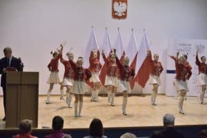 Na scenie stoją dziewczyny w czerwono-białych strojach. W rękach trzymają pałeczki mażoretkowe, a niektóre z nich mają jedną nogę podniesioną. Po lewej stronie za pulpitem stoi mężczyzna.