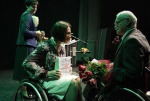 Kobieta na wózku inwalidzkim jest pochylona do przodu, a na kolanach trzyma torbę prezentową. Naprzeciwko niej jest mężczyzna na wózku inwalidzkim, który trzyma bukiet kwiatów.