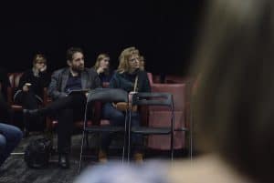 Osoby na widowni kierują wzrok w prawą stronę. Przed nimi stoją dwa puste krzesła. Jedna kobieta została sfotografowana siedząc tyłem.