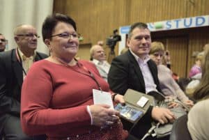 Osoby siedzą na widowni. Kobieta uśmiecha się a w ręce trzyma telefon. Obok mężczyzna patrzy wprost w obiektyw.