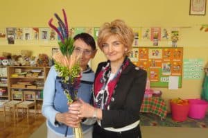 Dwie kobiety pozują do zdjęcia i uśmiechają się. Obie trzymają w rękach tę samą palme. Palma jest kolorowa.