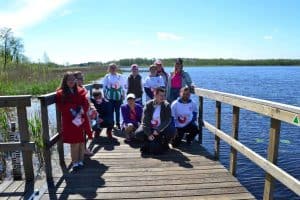 Grupa jedenastu uczestników, w tym cztery osoby w białych koszulkach z logiem Kilometry Dobra stoją na drewnianym pomoście i pozuje do zdjęcia na tle jeziora.