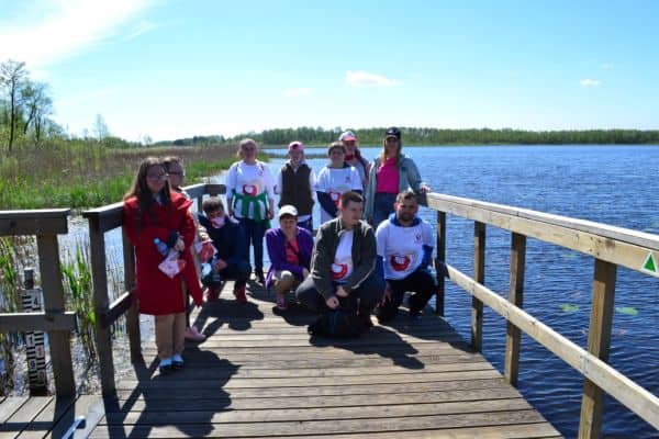 Grupa jedenastu uczestników, w tym cztery osoby w białych koszulkach z logiem Kilometry Dobra stoją na drewnianym pomoście i pozuje do zdjęcia na tle jeziora.