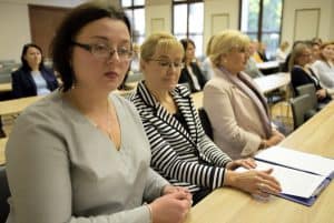 ludzie siedzą przy stołach. Ich twarze skierowane są na wprost. Na stole jest teczka z papierami.