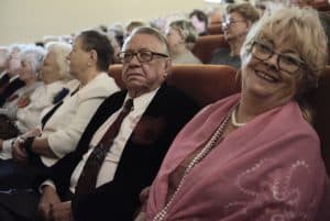 Mężczyźni i kobiety siedzą na fotelach. Kobieta patrzy wprost w obiektyw i uśmiecha się. Mężczyzna obok jest zwrócony w stronę obiektywu i patrzy poza niego.