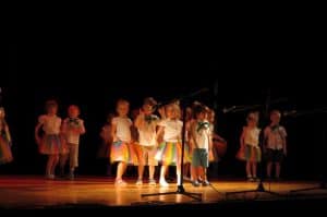 Dzieci w kolorowych spódniczkach i spodenkach stoją na scenie w grupie.