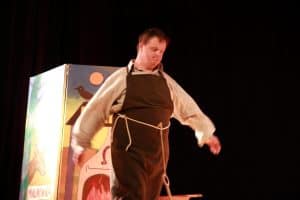 Na scenie stoi mężczyzna. Jest on przebrany za kowala. Za nim stoi scenografia z piecem.