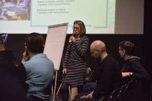 Kobieta stoi obok białej tablicy, trzymając w ręku mikrofon. W tle wyświetla się prezentacja. Dwóch mężczyzn z widowni patrzy w jej kierunku, zaś jeden pochyla głowę.