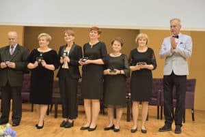 Grupa osób na scenie pozuje do zdjęcia. Sześć osób trzyma statuetki i stoi na scenie. Po prawej stronie mężczyzna wykonuje gest klaskania.