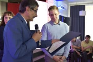 Mężczyzna trzyma mikforon i dyplom w dłoni. Za nim stoi młody mężczyzna, którego twarz jest zwrócona w kierunku dyplomu.