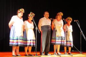 Ludzie w strojach ludowych stoją na scenie. Przed nimi są stojace mikrofony.