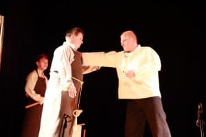 Na scenie stoją mężczyźni. Mężczyzna po prawej stronie trzyma wyprostowaną rękę na mężczyźnie po lewej stronie.