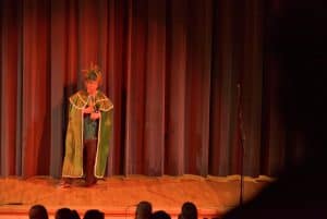 Mężczyzna stoi na scenie w zielonej pelerynie. W tle jest widownia.