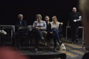 Kobieta trzyma mikrofon przy ustach i siedzi pośród trzech innych osób. Obok niej są dwa wolne krzesła. W oddali idzie mężczyzna ze złamaną ręką.