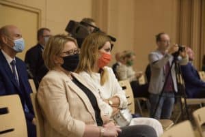 osoby siedzą na widowni. Po prawej stronie stoi mężczyzna z aparatem na statywie. Za nim jest mężczyzna, który trzyma kamerę.