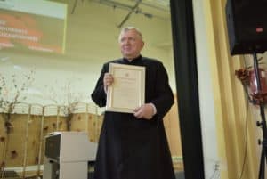 Ksiądz stoi na środku pomieszczenia. Ksiądz trzyma w rękach dyplom w ramce. Za nim na ścianie wyświetlany jest slajd.