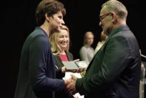 Kobieta i mężczyzna stoją naprzeciwko siebie a ręce mają złączone w uścisku. Kobieta w drugiej ręce trzyma medal. Oboje są bokiem do obiektywu.