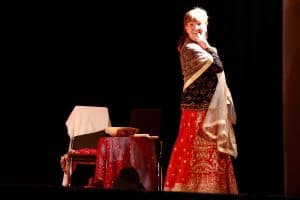 Kobieta po prawej stronie stoi w czerwonej spódnicy i szalu. Głowę ma skierowaną w stronę obiektywu. Za nią stoi krzesło i stolik.