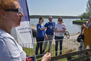 Trzy osoby stoją za barierką. Za nimi jest woda. Po lewej stronie jest kobieta w okularach, która jest bokiem do obiektywu.