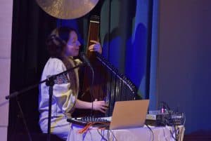 Kobieta jest na scenie. Dłonie ma położone na harfie elektrycznej. Przed nią jest stół z laptopem.